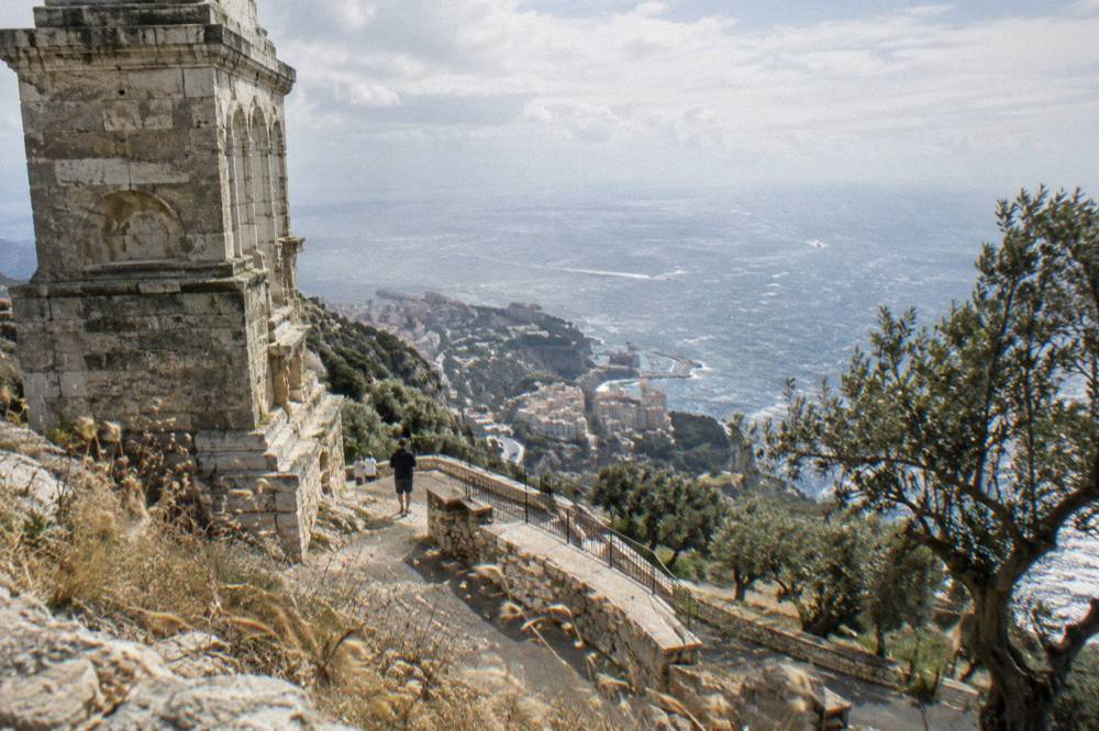 A stone monument terrace above a Mediterranean village with coastline in the distance.