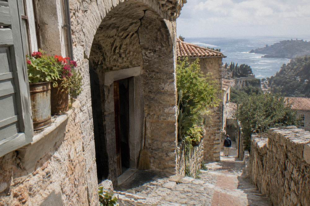 A narrow medieval village street with stone buildings and potted plants overlooking the sea.