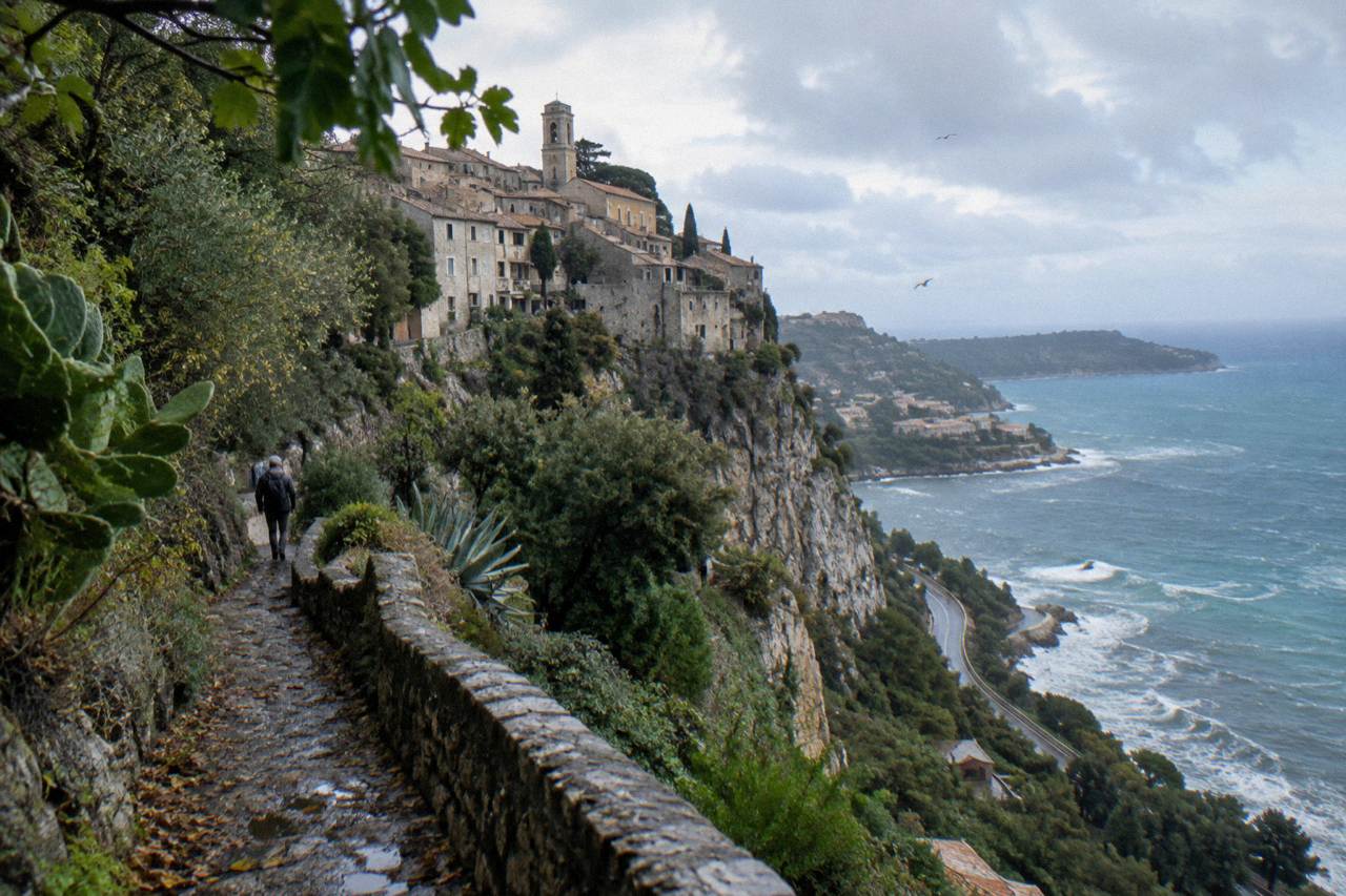 Stone village buildings on a cliff above the Mediterranean with a coastal road below