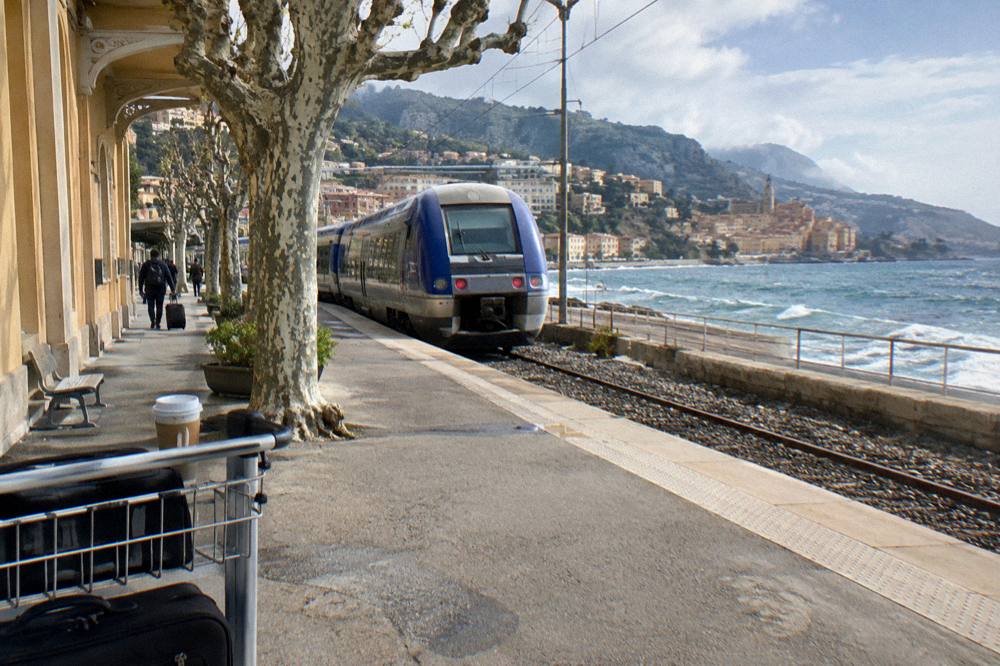 Blue commuter train at a platform beside a Mediterranean coastal station