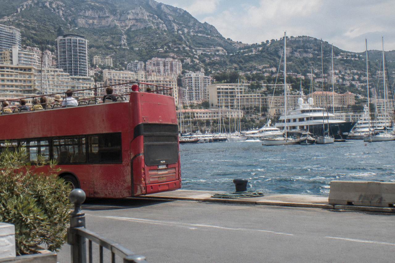 Open-top sightseeing bus parked beside Monaco harbor with hills and buildings behind