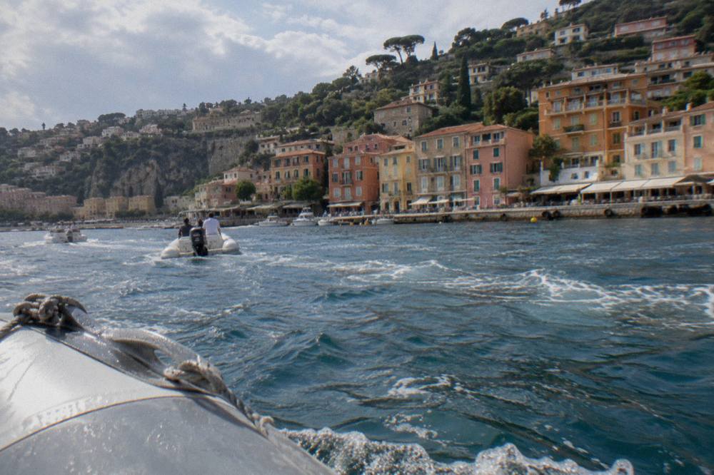 Tender boats crossing a calm blue bay toward a village waterfront with pastel buildings
