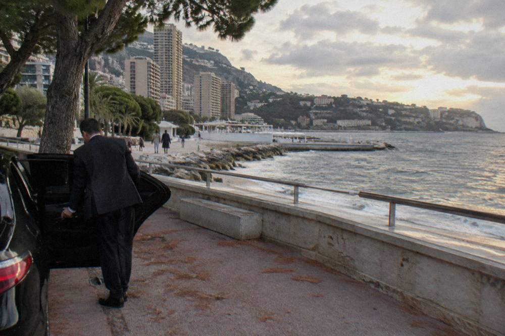 A chauffeur opening the rear door of a black sedan on a seaside promenade