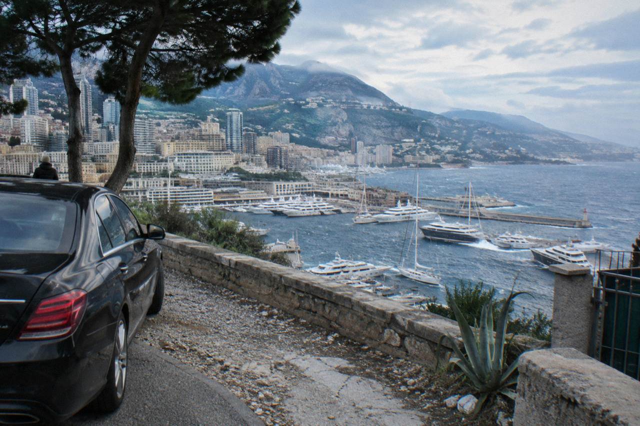 A luxury black sedan parked on a coastal road above Monaco harbor with hillside buildings behind it