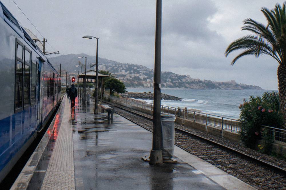 Railway platform beside the Mediterranean with a blue regional train and coastal hills beyond